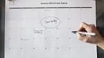 A printed January 2020 calendar posted on a on the wall with a hand noting something on it.
