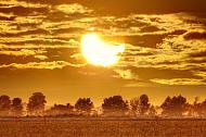 Picture with fields in the foreground and a crescent shaped Sun with Moon in front of it. Cloudy skies in the background.