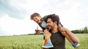 Photo of a little boy sitting on his fathers shoulders, out on a big field, laughing and holding a small plane in one hand.