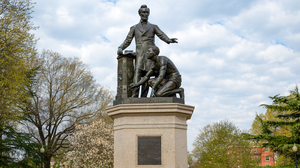 Statue of Abraham Lincoln standing next to a kneeling African American man, with broken shackles on his wrists, holding a document in his hand, with green trees in the background.