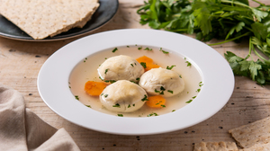 Picture of a bowl of traditional Jewish matzah ball soup and a plate with flat bread in the background, on a wooden table.