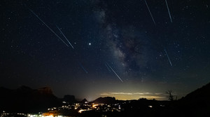 Dark sky with stars and several meteors over a landscape with city lights.