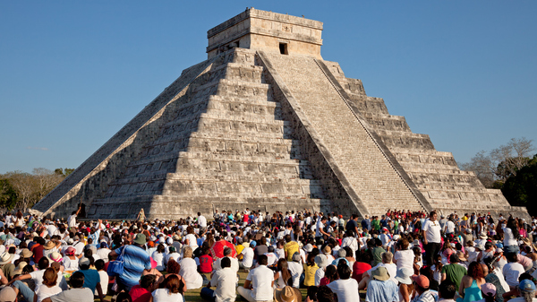 A large crowd gathered at a massive stepped structure made of light-colored stone, with a staircase running up its center under a clear sky.