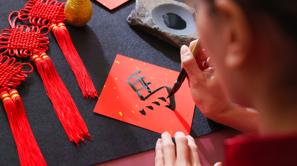 Top view of a woman writing a Chinese character meaning "horse" on a red sheet of paper with a brush, with red and golden ornaments lying next to the paper.