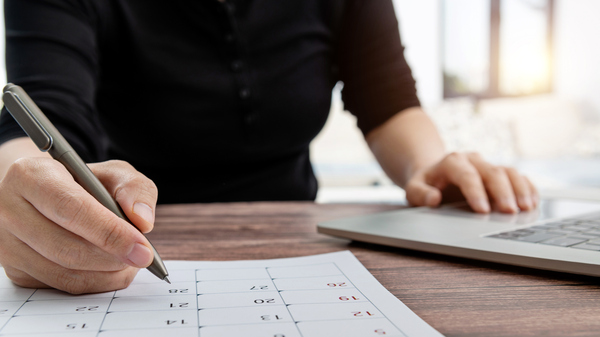A woman works on her computer while marking dates on a calendar with a pen.