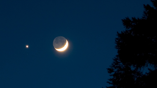 Crescent Moon with dark blue sky in the background. The unlit part of the Moon has a dim glow. Bright star shines to the left of the Moon.
