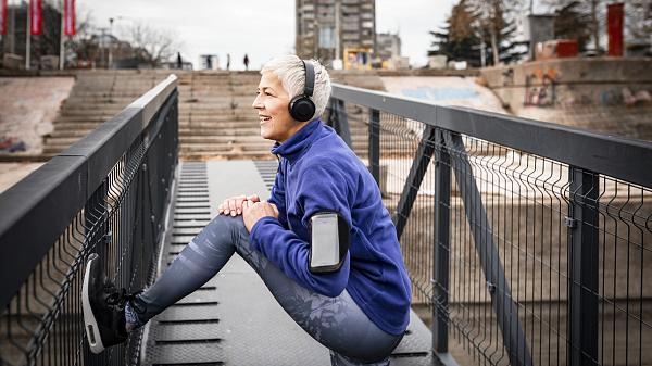 Grey haired senior woman in sports clothing stretching on a bridge and listening to music.