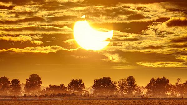 Picture with fields in the foreground and a crescent shaped Sun with Moon in front of it. Cloudy skies in the background.