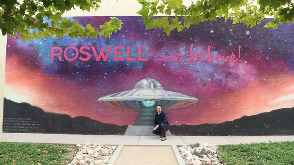Man posing in front of a mural showing a UFO and the words "Roswell ...we believe."