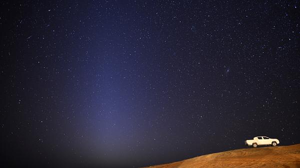 Starry night sky in the background with desert in the foreground. White truck on the side and a triangular glow of zodiacal lights in the sky.