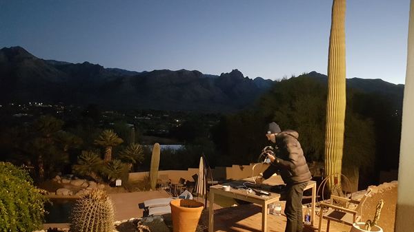 Man standing at a table outside at dawn, tidying up cables, with cacti, palm trees, and a mountain range in the background.