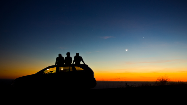 Silhouettes of three people sitting on a car roof looking at a twilit sky.
