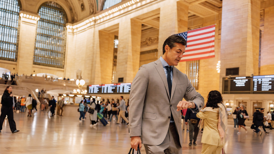 Business man in a suit glancing at his watch as he walks through a busy train station with more people and an American flag in the background.