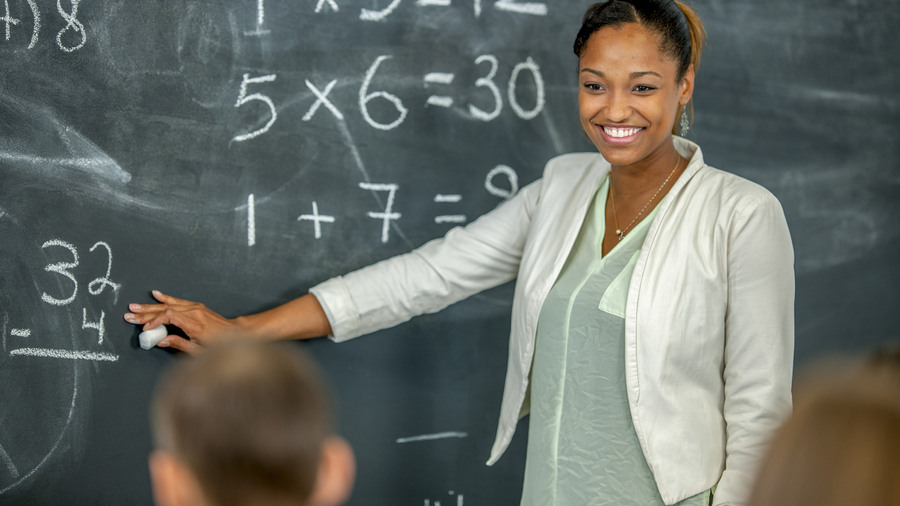 A smiling black woman pointing to a blackboard with math equations.