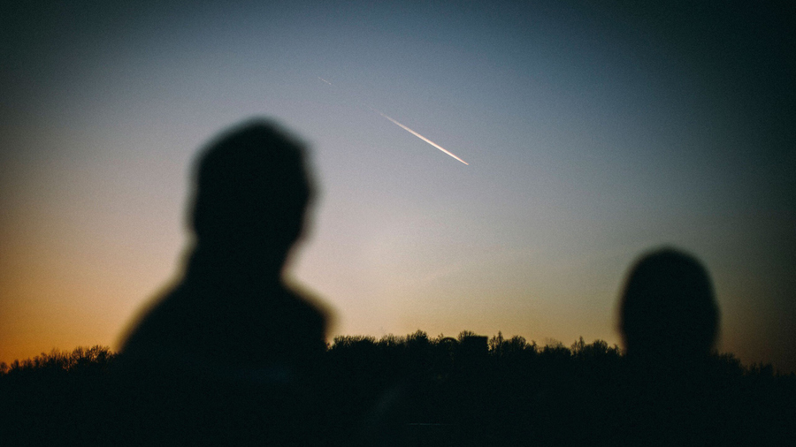 Silhouettes of two people watching a bright meteor streaking across the sky above a forested horizon.