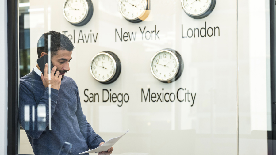 Man on phone with document, standing by world clocks for several world cities.