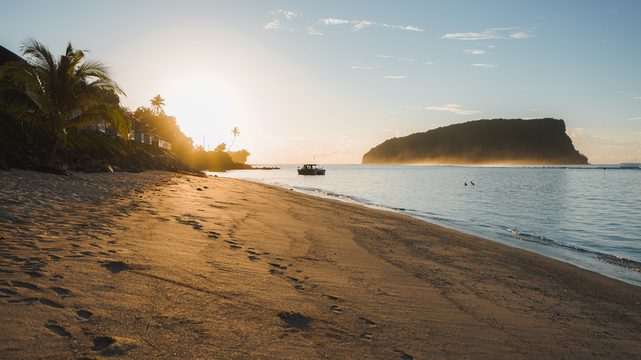 Sunset over a tropical beach with footprints, a boat, and a misty island in the distance.