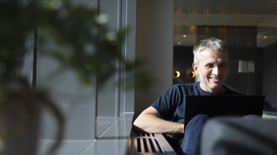 Man sitting by window looking at computer and smiling.