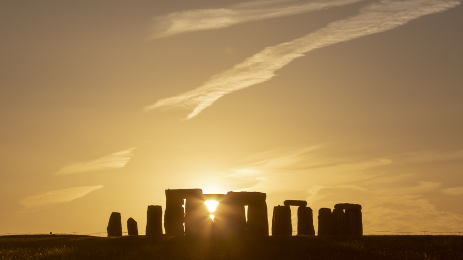 The Sun is rising behind a silhouette of stone pillars, casting a warm golden light across the sky and clouds.