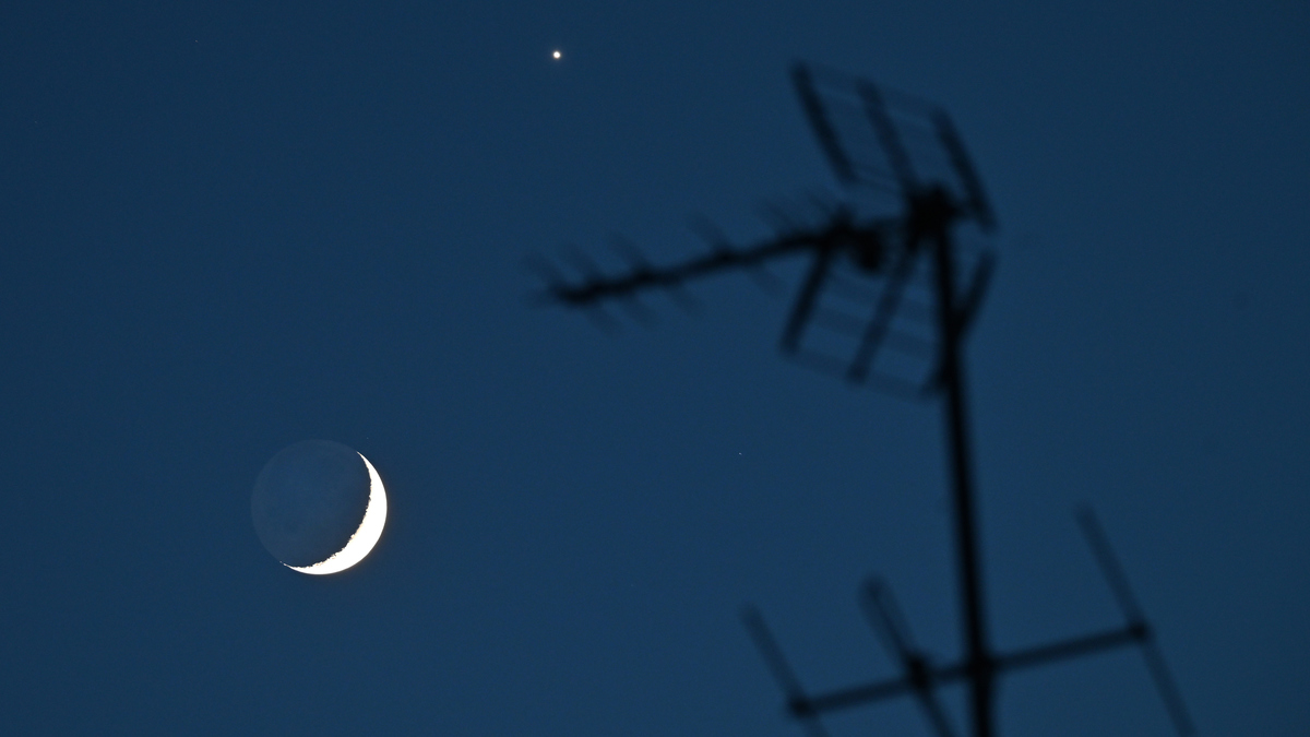 Crescent Moon and bright Venus in a dark blue sky, with the silhouette of a rooftop antenna in the foreground.