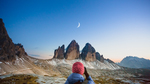 A woman hiker taking a picture of the Crescent Moon over three pointy mountains: Tre Cime in the Dolomites , Italy.