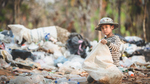 Small boy kneeling on a waste dump holding a large sack and collecting garbage.