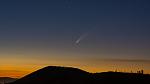 Comet in the dark sky over a mountain with a setting/rising Sun in the background.