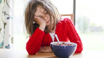 A child in a red jumper leaning their head in their hand and trying to eat a bowl of cereal.