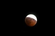 A partially eclipsed Moon in the night sky. A little over half of the Moon is covered by a reddish shade. 
