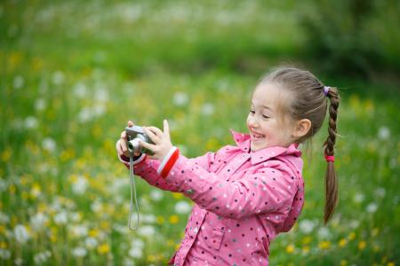 Little smiling and curious girl photographing with her camera exploring nature and standing in a dandelion meadow.