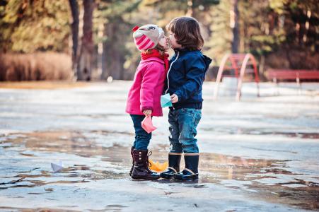 Toddler boy and girl kisses on a walk in spring puddle with paper boats in hands.