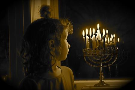 Ei ung jente ser på en delvis tent hanukkia, en niarmet lysestake som brukes under den jødiske hanukka-feiringen, som står på vinduskarmen. Det er mørkt utenfor vinduet.