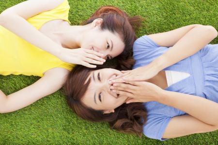 Two sisters whispering to each other while lying on grass.