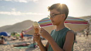 Boy eating ice cream in the Sun on a beach full of parasols and people.