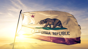 California state flag, flying in the wind in front of a setting sun. The flag shows a single red star in the canton, a red stripe along the bottom, and a California grizzly bear atop a mound of green grass defacing a white field.
