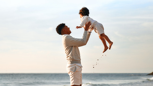 Father lifting young child into the air while smiling on a beach, with the ocean and sky in the background.