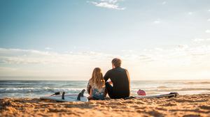 Father and daughter sitting on the beach with surfboards, looking out over the ocean.