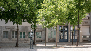 Courtyard with trees, a metal statue of a man, and a stone building in the background.
