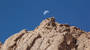 A Third Quarter Moon against the blue day sky over a sandy colored rock formation with soft eroded edges.