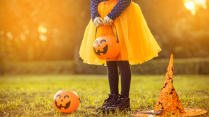 Child in an orange Halloween tutu holding a pumpkin bucket, with another bucket and a witch hat on the grass.