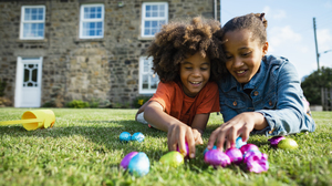 A young boy and his older sister lying on the grass, their house in the background, smiling and holding colorful Easter eggs.