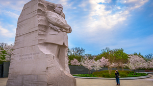A man is taking a photo of a large statue of Martin Luther King, with blossoming trees in the background.