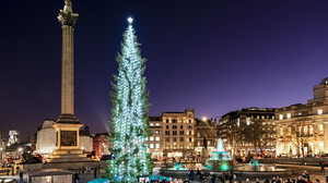 A big Christmas tree with bright lights standing in the middle of Trafalgar square at night. A choir is standing in front of the tree, singing, and people have gathered around them.