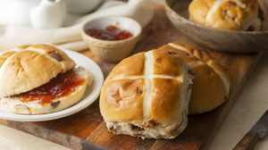 Picture of cross buns lying on a kitchen counter. One of the buns has been sliced in two and spread jam on.