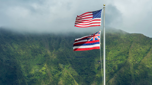 The Hawaiian and American flags flying high together in front of a mountain covered in greenery.