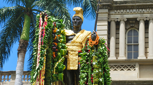 Statue of King Kamehameha decorated with colorful flower garlands with palm trees and blue sky in the background.