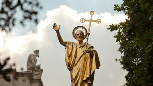 Golden statue of St. Paul raising his hand and holding a cross againts a blue sky, some greenery and the top of an old stone building with a statue sitting on top, in the background.