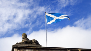 Scottish blue and white flag waving on top of an old building, next to stone statue, with blue sky and a few white clouds in the background.