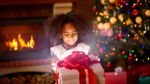 Little african american girl opening a Christmas present in front of the Christmas tree and fireplace.