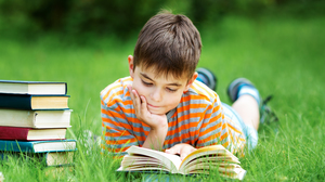 Young boy lying on the green grass, reading a book.